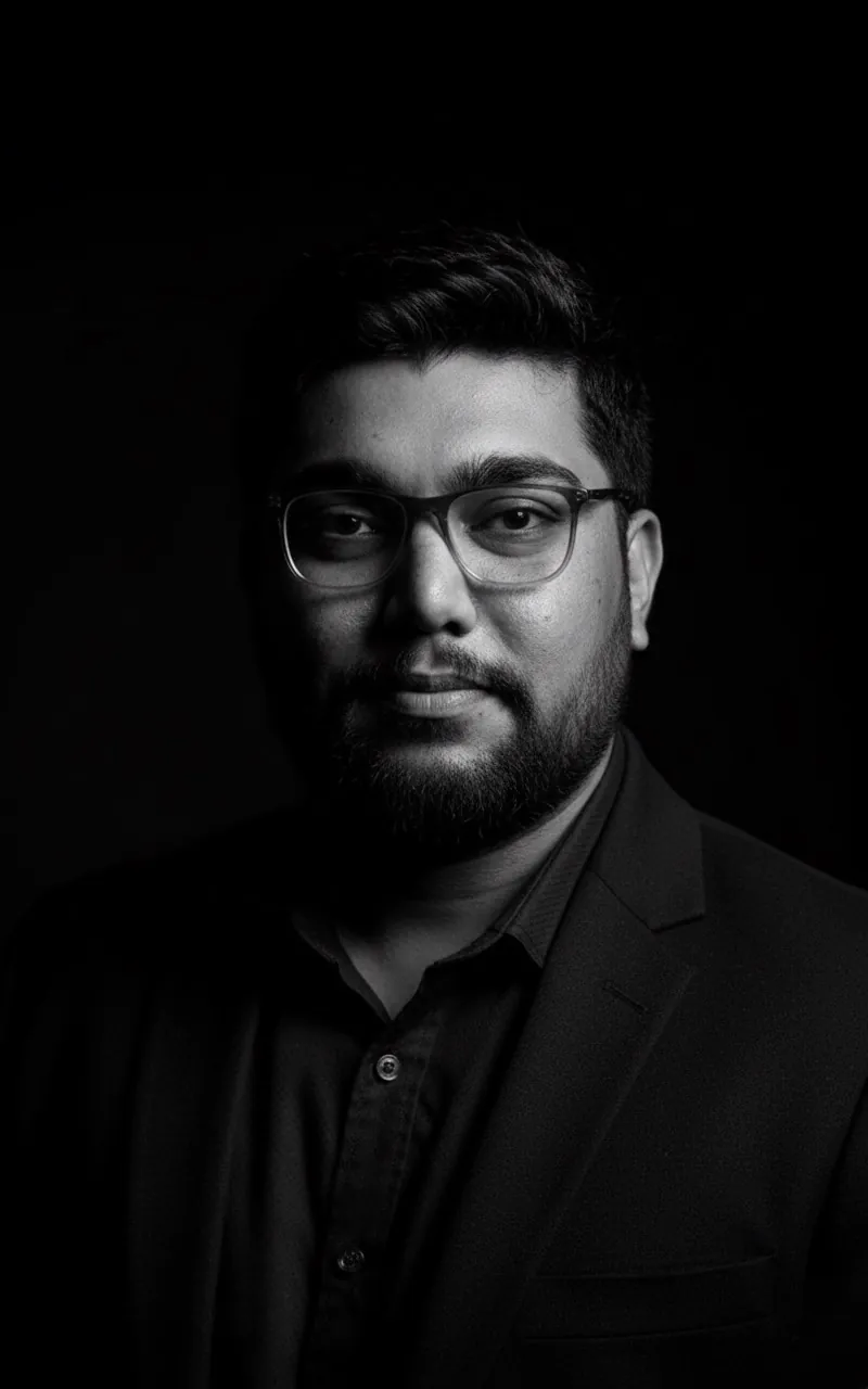 Black and white studio portrait of Aneel, wildlife photographer, wearing glasses and a dark jacket
